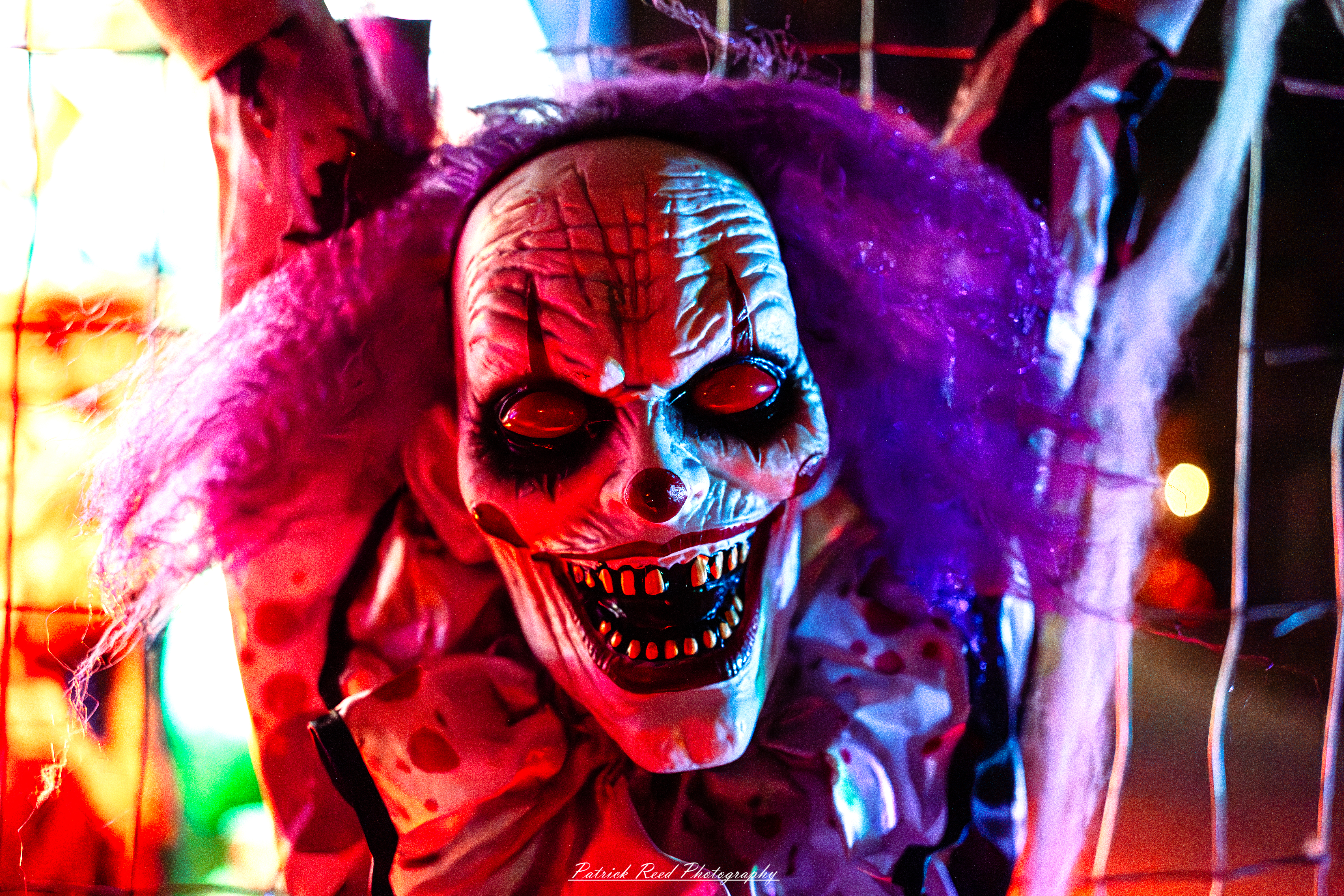 A chilling close-up portrait of a nightmare clown with ghastly makeup and a sinister grin. The clown’s face is painted in smudged white, with dark, hollow eyes that seem to stare right through the viewer. Its lips curl into a twisted smile, revealing jagged, decaying teeth. Dark shadows emphasize the deep wrinkles and eerie contours of its face, and faint streaks of red paint drip down from exaggerated cheekbones. The entire image is dimly lit, with an unsettling, smoky haze in the background, making the clown’s gaze feel even more menacing. Taken on Tilson Street in Romeo Michigan on Halloween.